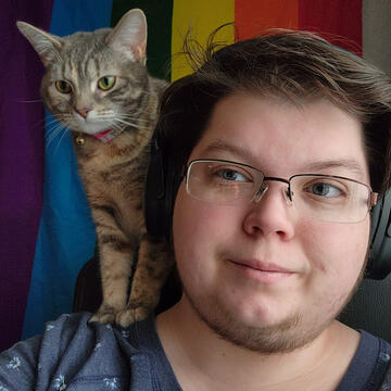 A selfie of Nix Song, with their grey and white cat sitting on his shoulder in a way that makes it look like she is "supervising" Nix. There is a rainbow flag in the background.